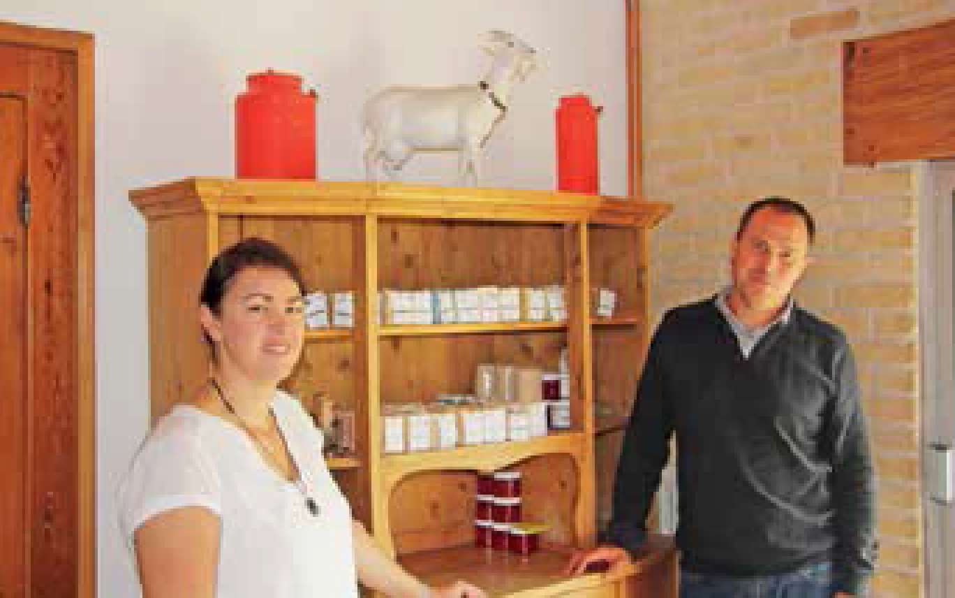 Claudia et Sylvain Janquin devant un présentoir exposant leurs savons. La chèvre et les deux pots sur le meuble symbolisent l’emploi du lait de chèvre dans la fabrication de leurs savons.